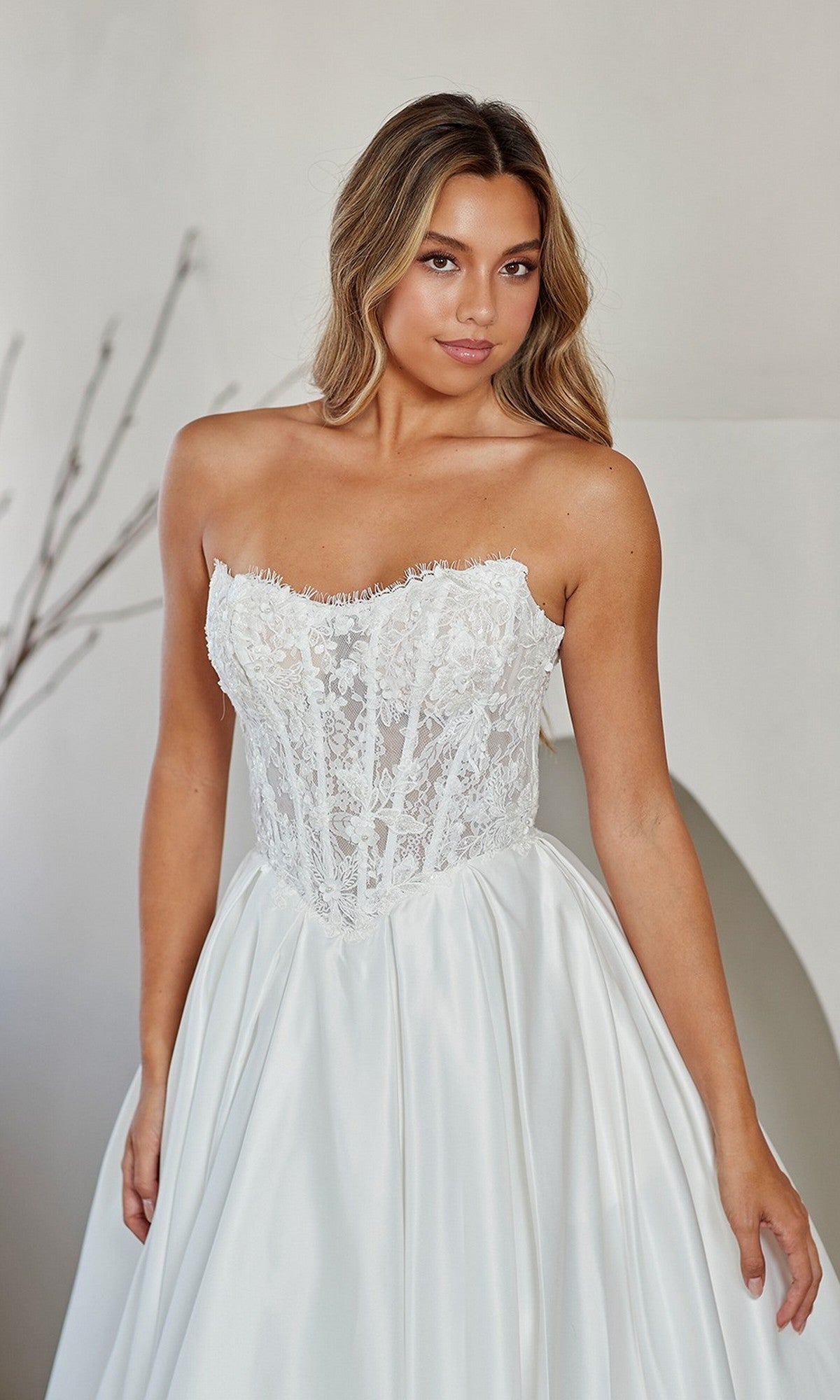 A woman wearing the Amelia Couture 2089 long prom dress, featuring a strapless white lace corset bodice and satin skirt, stands indoors against a neutral background.